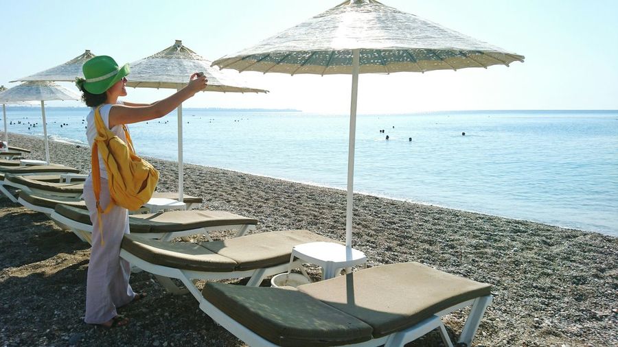 Woman photographing with smart phone while standing by parasols and lounge chairs at beach