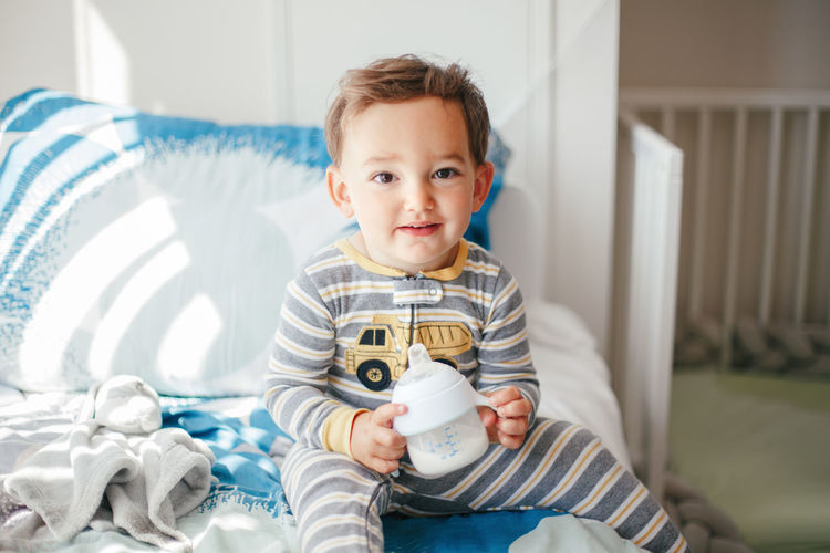 Cute adorable caucasian kid boy sitting on bed drinking milk from kids bottle. healthy eating 
