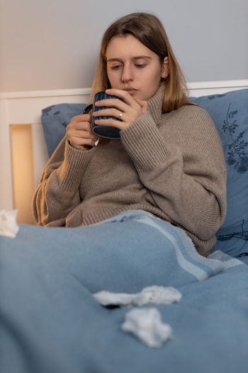 Cute girl in sweater sitting in bed under blue blanket and drink hot beverage. woman feeling sick.