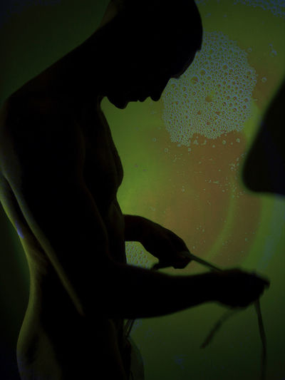 Silhouette of a young man nude in the dark with light and shapes projections on a wall, digital 