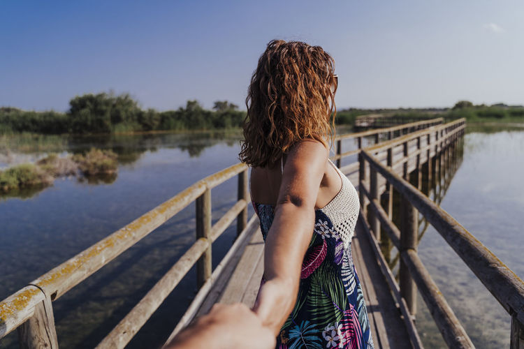 Woman holding hand of boyfriend while standing on footbridge against sky
