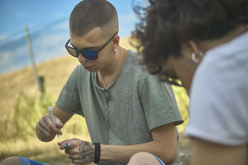 Young man holding marijuana joint while sitting by friend outdoors