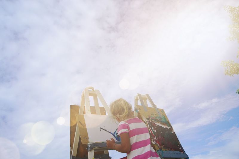 Rear view of girl making painting on canvas against cloudy sky