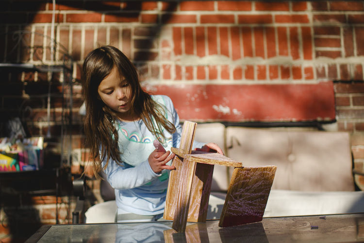 A focused child concentrates on building with wood against brick wall