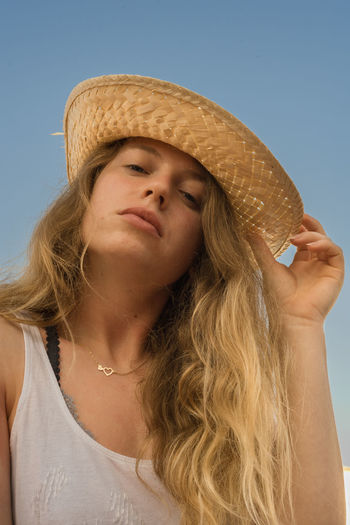 Low angle portrait of young woman wearing hat against clear sky