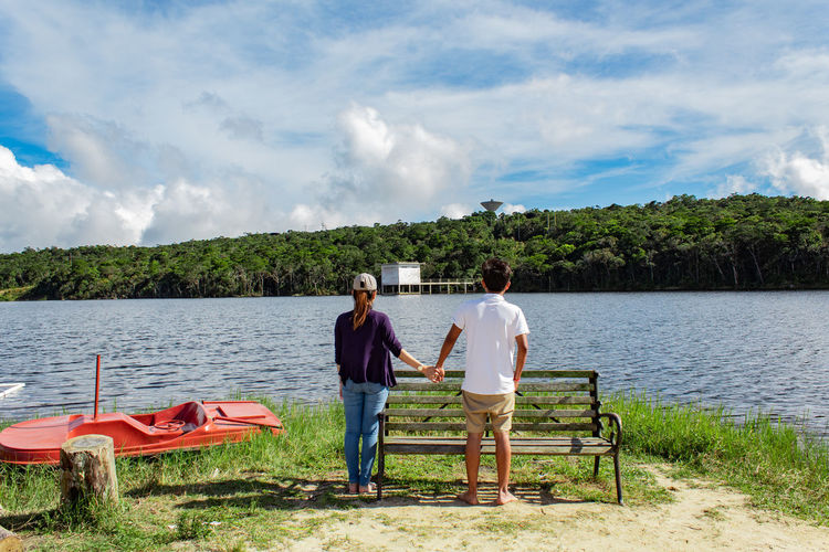 Rear view of couple holding hands while standing on field by lake against cloudy sky during sunny day