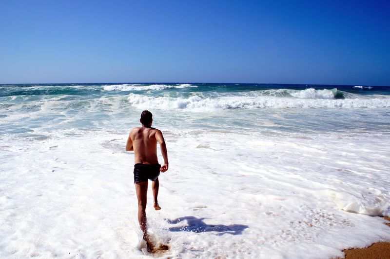 Rear view of shirtless man walking on beach against clear sky