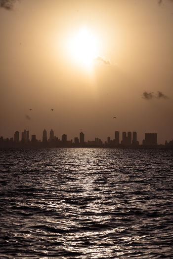 Silhouette buildings by sea against sky during sunset