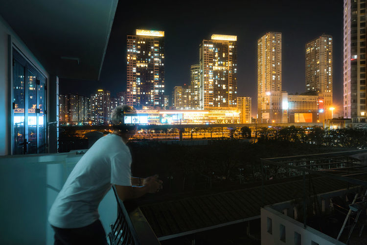 Man looking at illuminated buildings while standing in balcony at night