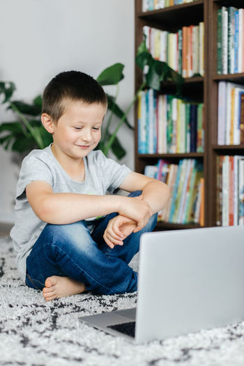 A schoolboy boy sits on the floor in front of a laptop and watches educational videos. distance