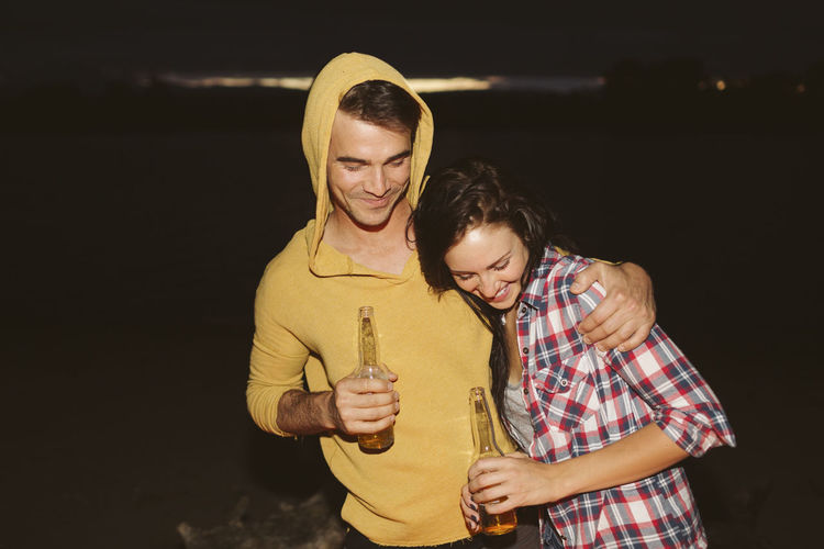 Happy friends with arm around holding bottles while standing by river at night