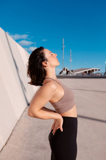 Side view of fit female in sportswear standing back bending while resting with eyes closed on street against blurred background during training on sunny summer day