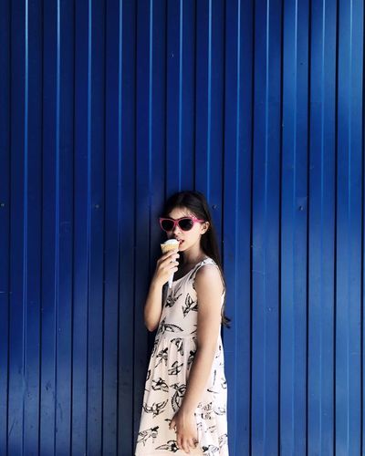 Portrait of girl eating ice cream while standing against blue corrugated iron