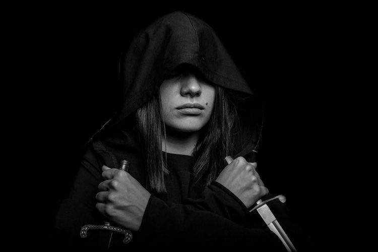 Young woman wearing hood holding swords against black background
