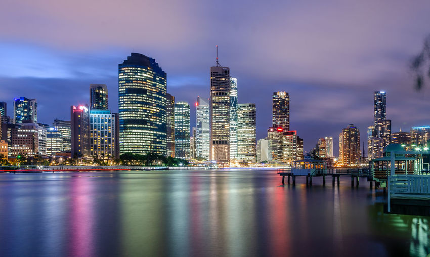 Illuminated modern buildings by bay against sky
