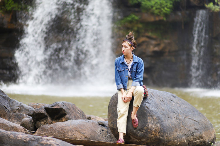 Full length of woman sitting on rock against waterfall