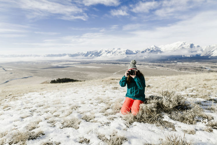 Female hiker photographing with camera while kneeling on snowy field against mountains