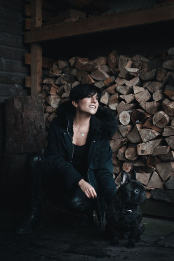Smiling woman with dog crouching against firewood