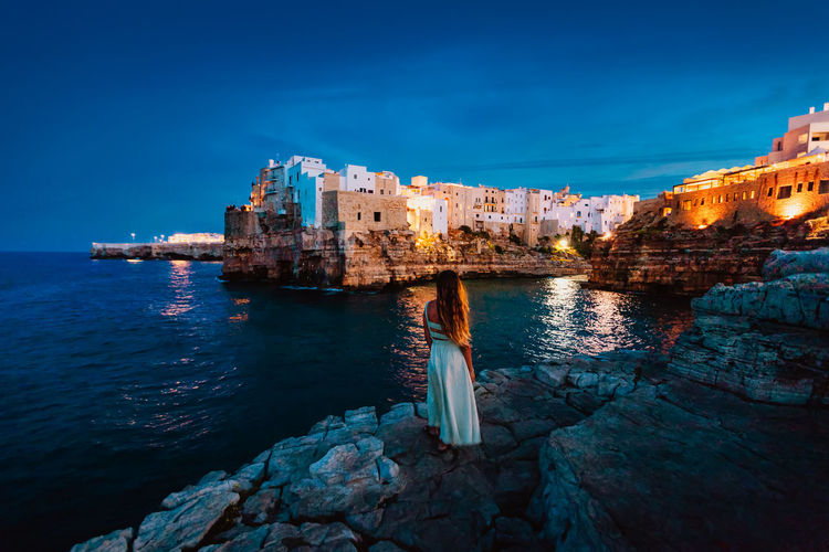 Young woman on the rocks in front of the apulian village of polignano a mare at sunset 