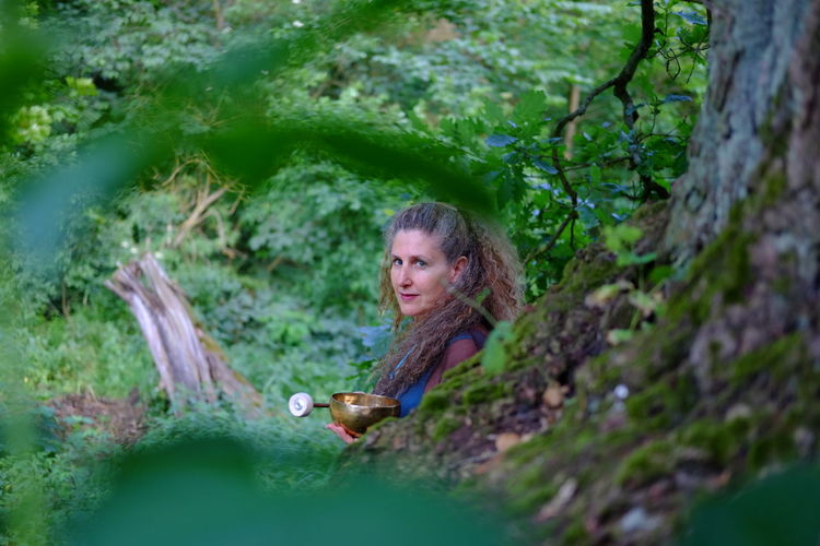 Portrait of mature woman holding singing bowl while sitting in forest