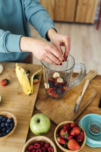 Woman putting fresh chopped fruits in blender at table in kitchen