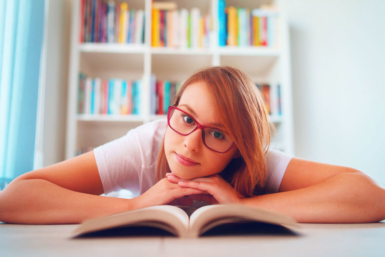 Portrait of beautiful woman wearing eyeglasses reading book lying on floor at home