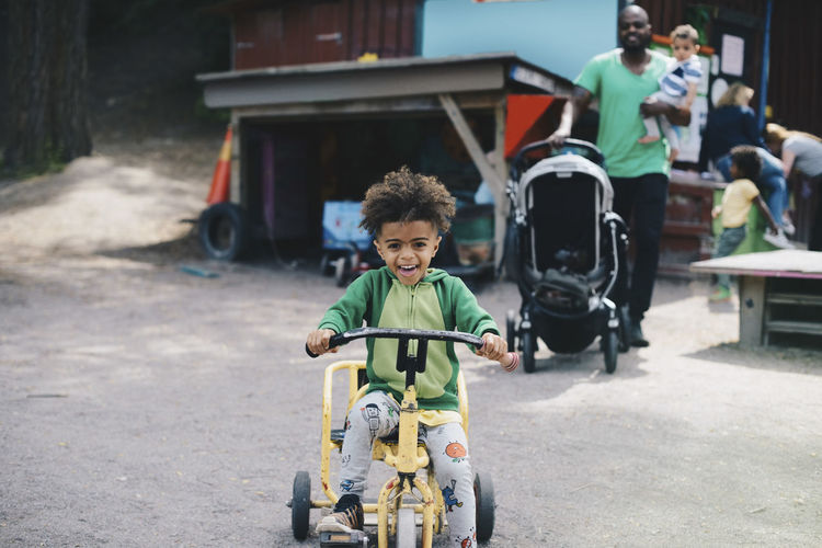 Happy boy riding tricycle while father and brother walking in background