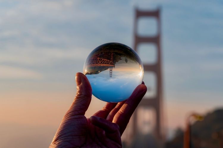 Close-up of hand holding crystal ball against golden gate bridge during sunset