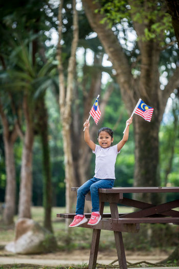 Portrait of smiling girl holding malaysian flag while sitting on bench at park