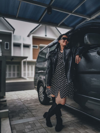 A girl wearing a polkadot dress, jacket, sun glasses and high boots standing outside the car