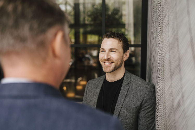 Smiling businessman looking at male professional discussing while standing by wall during seminar at convention center