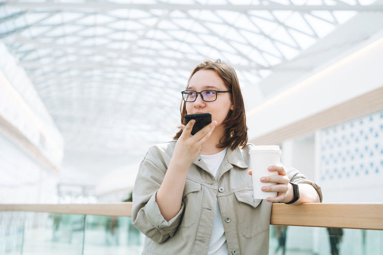 Young teenager girl in glasses using mobile phone sent voice message with coffee at shopping mall