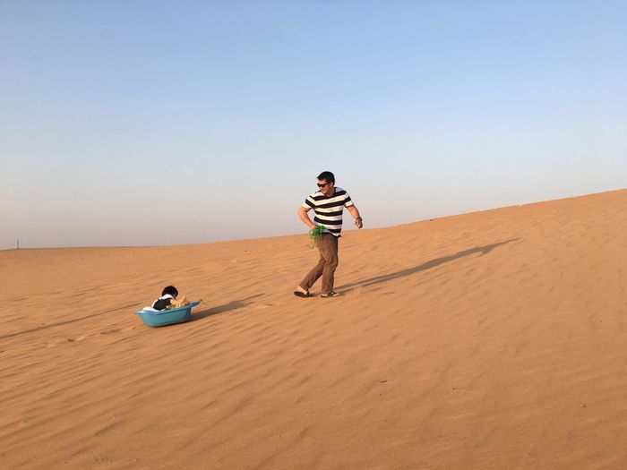 Father pulling son in small bathtub at desert against sky