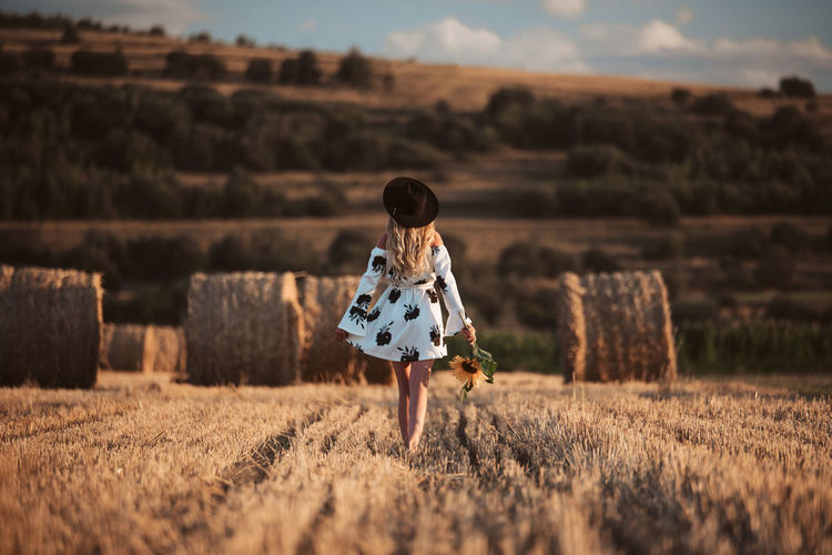 Woman in white dress walking in sunset field with hay bales, beautiful romantic girl with long hair
