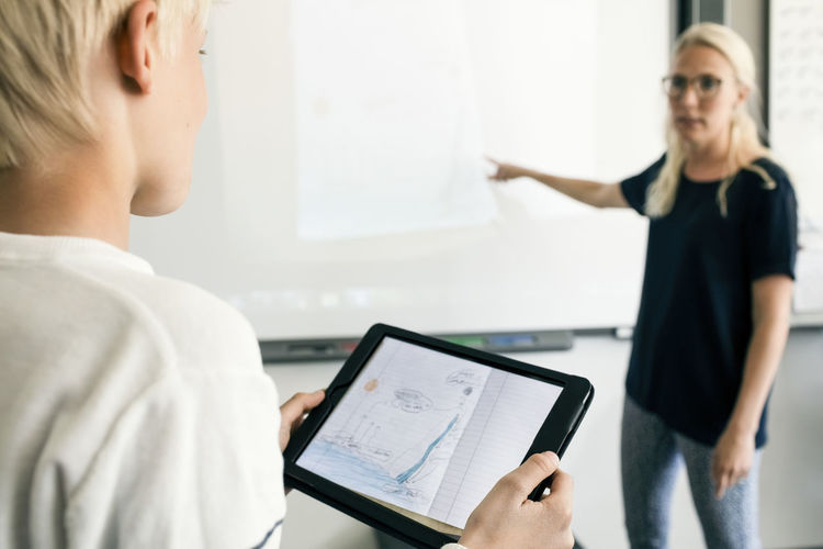 Cropped image of boy holding digital tablet with teacher explaining in classroom
