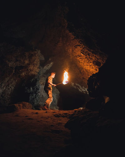 Side view of young male speleologist with flaming torch standing in dark narrow rocky cave while exploring subterranean environment
