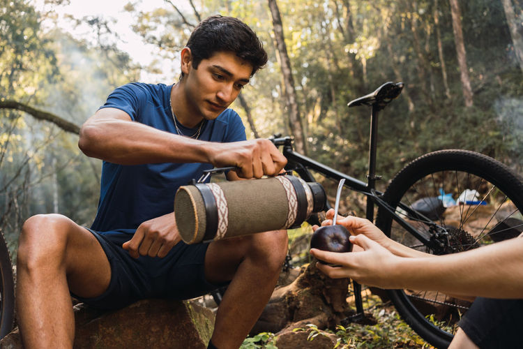 Serious ethnic male sitting on log and pouring hot drink from thermos into anonymous mate cup in hands of woman during hike on bicycles in woods