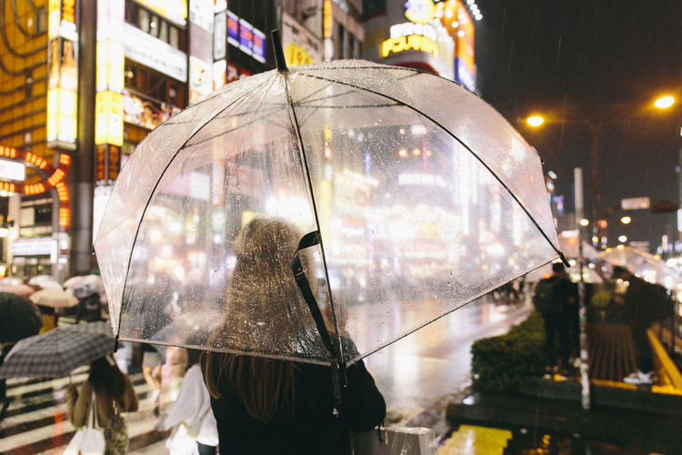 Rear view of woman with umbrella standing on road during rainy season in city at night
