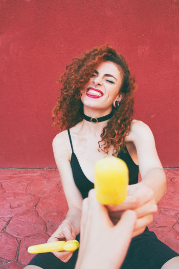 Cropped hand giving cheerful female friend popsicle against red wall