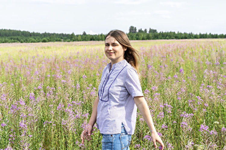 Young beautiful smiling blond woman in purple shirt walking in meadow among flowers fireweed, field