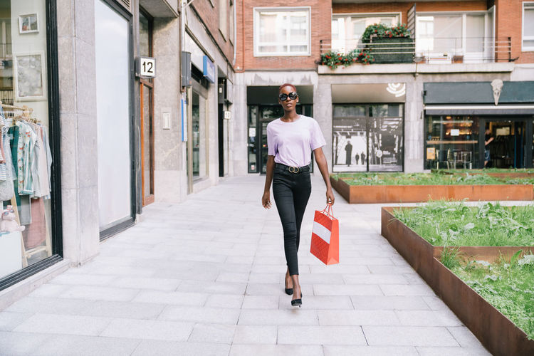 Full body of confident stylish young african american female shopaholic in trendy clothes and sunglasses with shopping bag in hand walking on street near fashion store