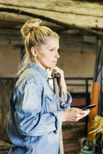 Thoughtful woman holding phone standing in barn