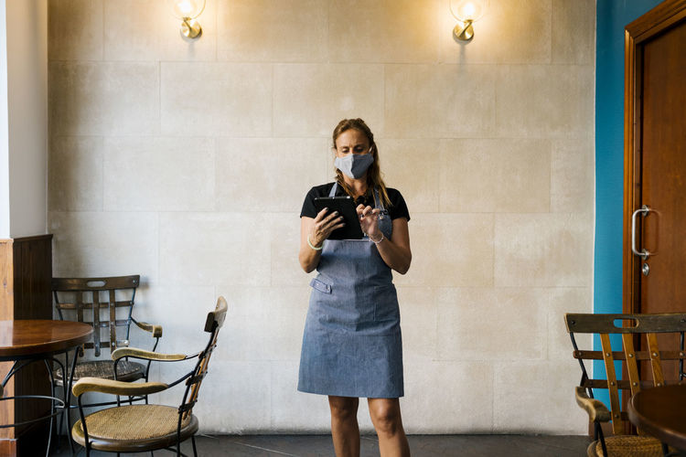 Female cafe owner wearing protective face mask while using digital tablet against wall