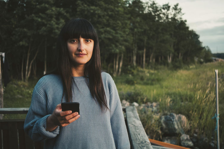 Portrait of smiling woman using mobile phone while standing by lake against sky