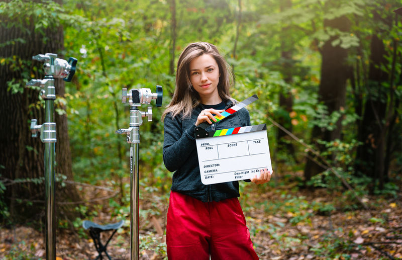 Portrait of young woman holding clapper board in forest. film slate