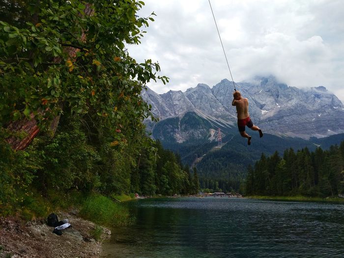 Full length of shirtless man jumping in lake from rope against mountains