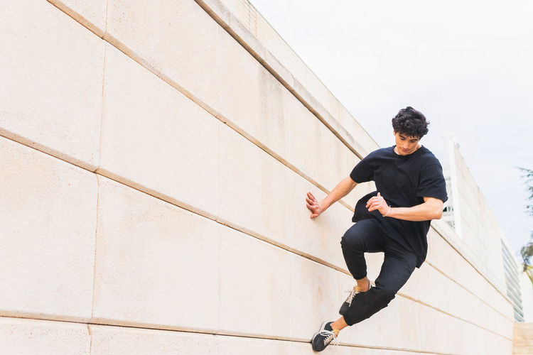 Side view of full length man in black outfit performing acrobatic parkour stunt while jumping above wall on building