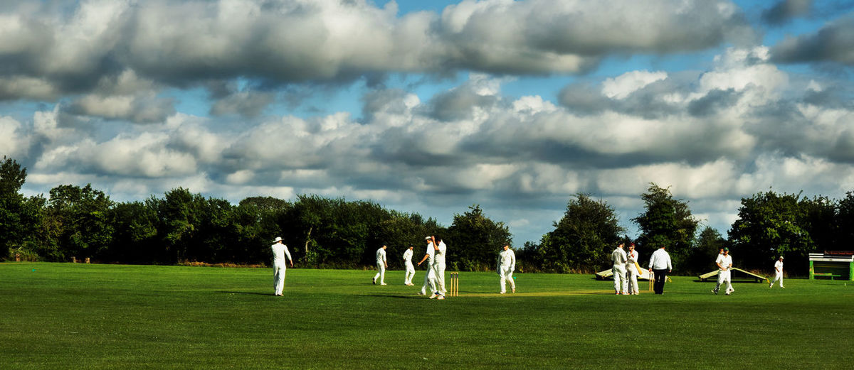 Panoramic view of people playing cricket on field against sky