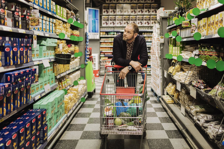 Thoughtful male customer leaning on shopping cart at supermarket