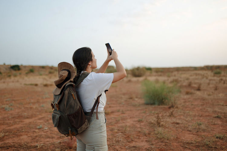 Back view of female traveler with backpack taking self portrait on smartphone while standing in desert area during trip on summer day